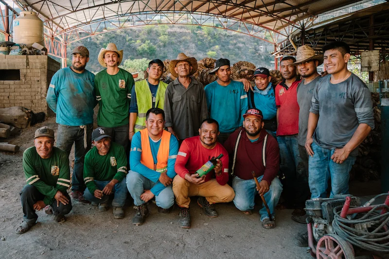 Group portrait of master mezcaleros and distillery workers at a traditional mezcal palenque in Oaxaca with harvested agave piñas in the background.