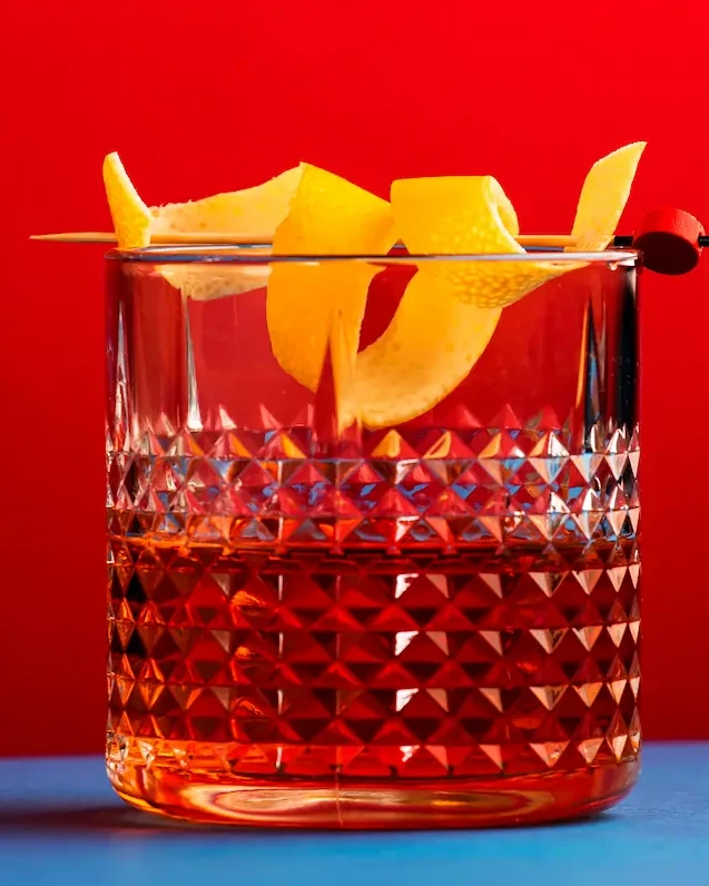 Cocktail served in a cut-glass rocks glass with an orange twist garnish against a bold red background.