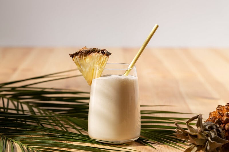 Cóctel tropical de mezcal con piña y coco servido en vaso highball con garnish de piña y pajita metálica.