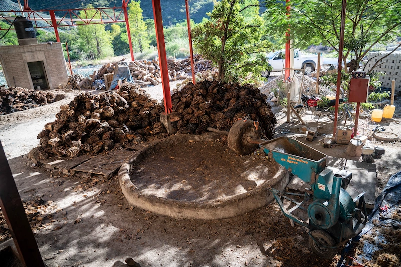 Mezcal Grinding Area with Stone Tahona and Agave Shredder Overview of a mezcal distillery setup showing piles of cooked agave piñas, a circular stone tahona mill, and a green mechanical shredder.