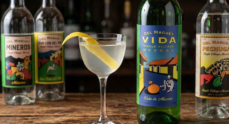 portrait for paciano stirred mezcal italicus cocktail in nick and nora glass with vida de muertos bottle and lemon twist garnish