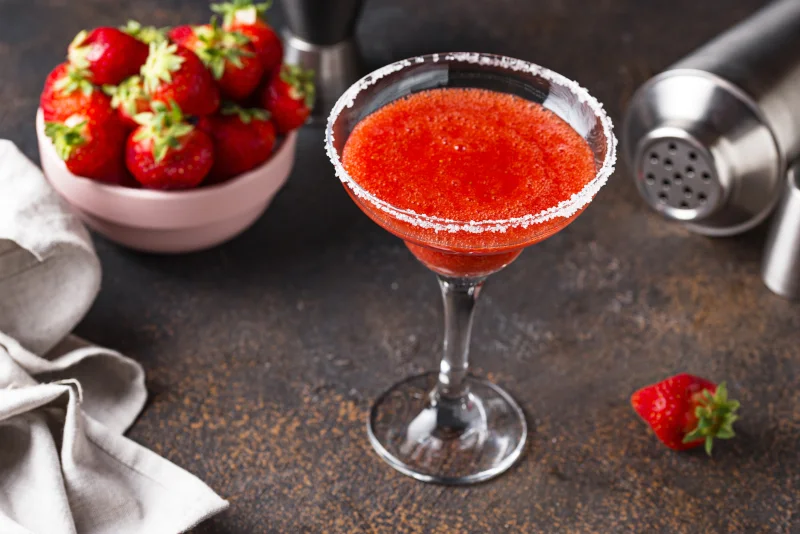 Fresas machacadas, lima y jarabe de agave en una coctelera junto a una botella de Del Maguey Vida Clásico