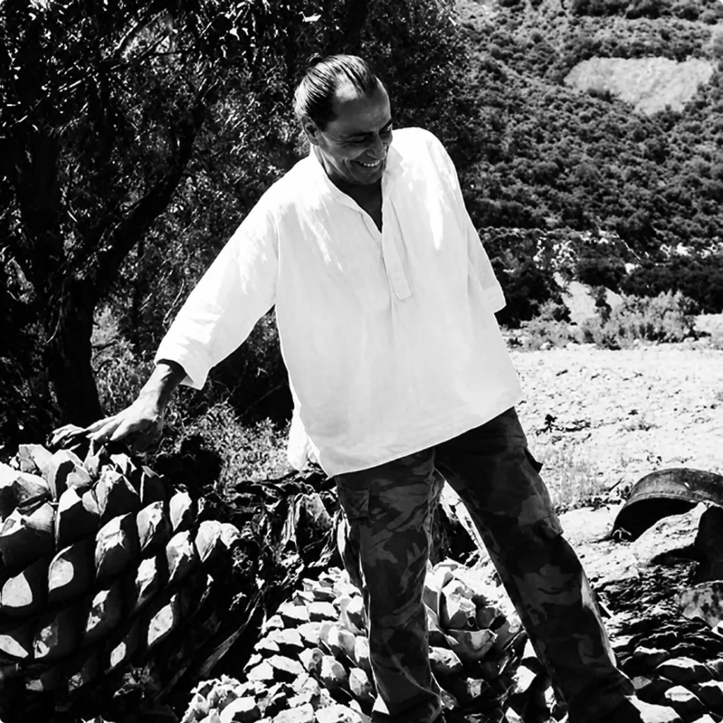 Black and white photograph of a man in a white shirt beside a pile of split agave pinas at a palenque